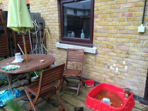 Patio, house wall with window to rear of photo. Wooden table and chairs, with parasol, in front left of photo. Small red plastic paddling pool front right.